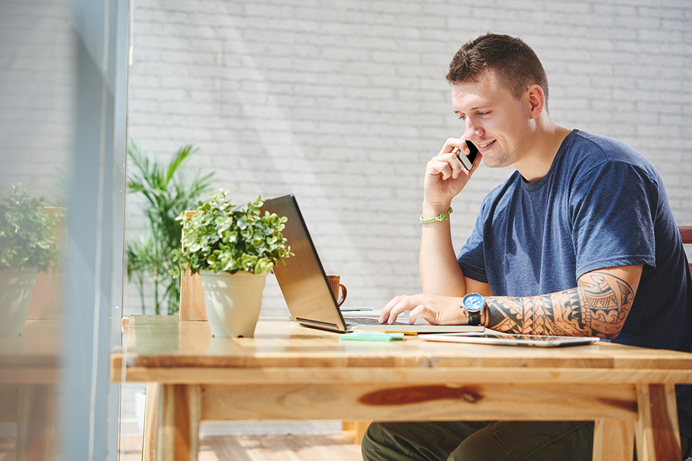 A young man working on the computer and calling someone on the phone.